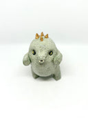 A small ceramic sculpture of a baby dinosaur with a textured light gray-green body, three gold horns on its head, and large yellow eyes, standing against a plain white background.