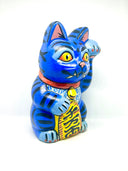 A hand-painted blue maneki-neko lucky cat figurine with black stripes, yellow eyes, pink inner ears, and a red collar, holding a yellow sign with black kanji characters, set against a plain white background.