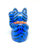 A hand-painted blue maneki-neko lucky cat figurine with black stripes, yellow eyes, pink inner ears, and a red collar, holding a yellow sign with black kanji characters, set against a plain white background.