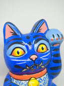 A hand-painted blue maneki-neko lucky cat figurine with black stripes, yellow eyes, pink inner ears, and a red collar, holding a yellow sign with black kanji characters, set against a plain white background.
