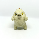 A small textured ceramic sculpture of a baby dinosaur with a light green-colored body, yellow eyes, and small gold spikes, standing against a plain white background.