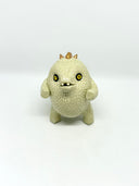 A small textured ceramic sculpture of a baby dinosaur with a light green-colored body, yellow eyes, and small gold spikes, standing against a plain white background.