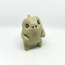 A small textured ceramic sculpture of a baby dinosaur with a light green-colored body, yellow eyes, and small gold spikes, standing against a plain white background.