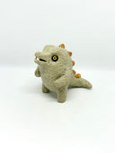 A small textured ceramic sculpture of a baby dinosaur with a light green-colored body, yellow eyes, and small gold spikes, standing against a plain white background.
