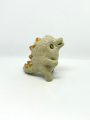 A small textured ceramic sculpture of a baby dinosaur with a light green-colored body, yellow eyes, and small gold spikes, standing against a plain white background.