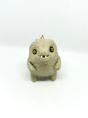A small textured ceramic sculpture of a baby dinosaur with a light green-colored body, yellow eyes, and small gold spikes, standing against a plain white background.