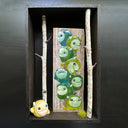 Open shadowbox artwork of a small yellow horned monster sculpt. It stands next to a vertical panel with many more monsters of the same kind painted on, mostly blue and greens. Birch branches are on either side.