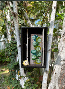Open shadowbox artwork of a small yellow horned monster sculpt. It stands next to a vertical panel with many more monsters of the same kind painted on, mostly blue and greens. Birch branches are on either side.