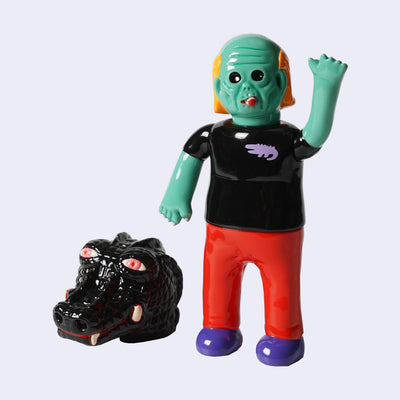 Vinyl figure of a green man, with sunken black eyes and wrinkled skin akin to a zombie. He smokes a cigar and wears a shirt with a small crocodile logo. Nearby is a large black crocodile head, with pink glazed over eyes.