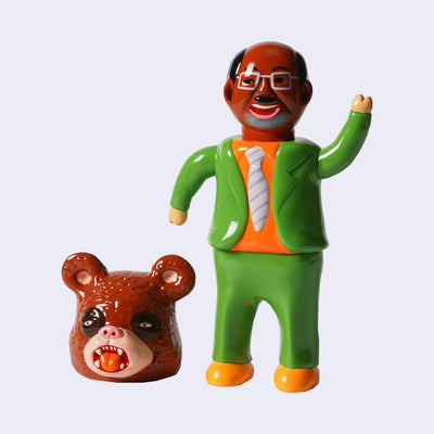 Vinyl doll of a tan business man, with a green suit, orange shirt and gray tie. He wears orange shoes and has bear paws for hands. Next to him is a large brown panda head with its mouth open.