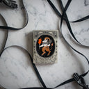 Small grey plaque with a small illustration in a carved out oval in the center. Drawing is of a goat demon with horns, a tail and holding a flaming torch and bundle of sticks.