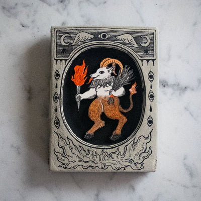 Small grey plaque with a small illustration in a carved out oval in the center. Drawing is of a goat demon with horns, a tail and holding a flaming torch and bundle of sticks.