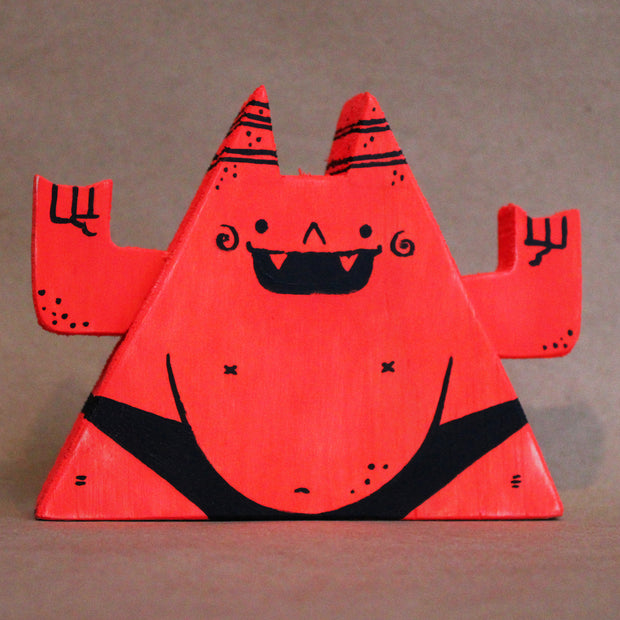 Wooden die cut sculpture of a red demon, shaped like a triangle with a very round belly. It wears only a black underwear and has its arms up at its side, making 2 "rock on" symbols.