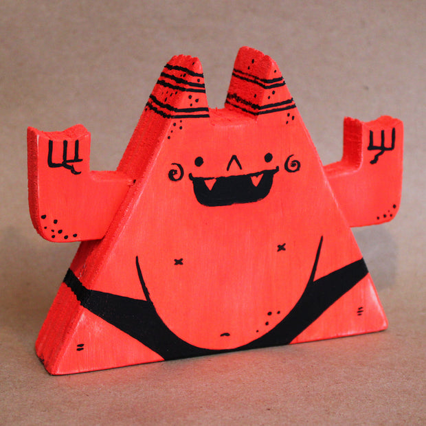 Wooden die cut sculpture of a red demon, shaped like a triangle with a very round belly. It wears only a black underwear and has its arms up at its side, making 2 "rock on" symbols.