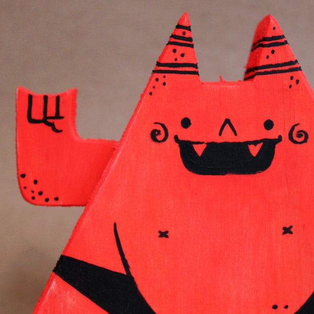 Wooden die cut sculpture of a red demon, shaped like a triangle with a very round belly. It wears only a black underwear and has its arms up at its side, making 2 "rock on" symbols.