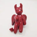 Ceramic sculpture of a red devil, with simplistic curved limbs and horns. It has a simple drawn on face and wings and a tail.