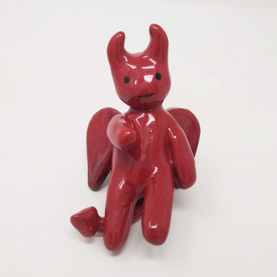 Ceramic sculpture of a red devil, with simplistic curved limbs and horns. It has a simple drawn on face and wings and a tail.