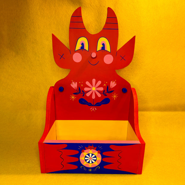 Wooden shelf with a large backing shaped like a cute, red devil with floral patterning on its chest.