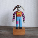 Painted whittled wooden sculpture of a cartoon style dog, standing up like a human and wearing a striped shirt and jeans. It has long, floppy ears and is half brown and half white.