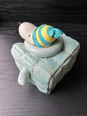 Ceramic sculpture of Snoopy wearing a puffy teal colored jacket and blue and yellow striped beanie. His body is shaped like a cube.