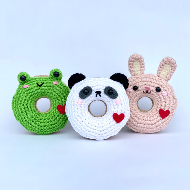 3 crocheted donuts fashioned like different animals, including a frog, a panda and a pink bunny. They each have small cute faces with pink cheeks and a red heart on their side.