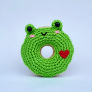 Crocheted donut fashioned like a frog, green with 2 slightly raised eyes. It has a cute smiling face and a red heart on its side.