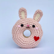 Crocheted donut fashioned like a pink bunny rabbit with raised ears. It has a cute smiling face and a red heart on its side.