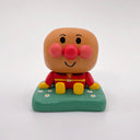 Wooden carving of a cute, round headed character with red cheeks and a nose sitting on a mound of grass.