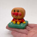 Wooden carving of a cute, round headed character with red cheeks and a nose sitting on a mound of grass.