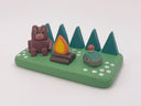 Camping-themed figure scene with a bear, campfire, and smiling rock with a mushroom growing atop it - all on a green base with trees and flowers.