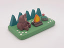 Camping-themed figure scene with a bear, campfire, and smiling rock with a mushroom growing atop it - all on a green base with trees and flowers.