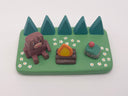 Camping-themed figure scene with a bear, campfire, and smiling rock with a mushroom growing atop it - all on a green base with trees and flowers.