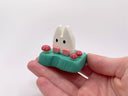 Small wooden sculpture of a white Chibi Totoro sitting on a green base with tiny mushroom around. Held in someone's hand.