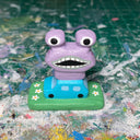 Small painted wooden sculpture of a tiny blue car on a painted green field with a large purple monster atop of it.