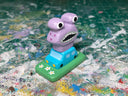 Small painted wooden sculpture of a tiny blue car on a painted green field with a large purple monster atop of it.