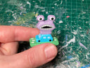 Small painted wooden sculpture of a tiny blue car on a painted green field with a large purple monster atop of it.