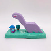 Purple dinosaur-like creature with a small house and tree on a green base against a white background