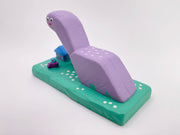 Purple dinosaur-like creature with a small house and tree on a green base against a white background