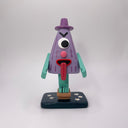 Sculpture of a triangular folklore character with one eye, a nose, its tongue sticking out of its mouth and standing on one leg, which wears a wooden sandal.