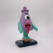 Sculpture of a triangular folklore character with one eye, a nose, its tongue sticking out of its mouth and standing on one leg, which wears a wooden sandal.