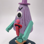 Sculpture of a triangular folklore character with one eye, a nose, its tongue sticking out of its mouth and standing on one leg, which wears a wooden sandal.