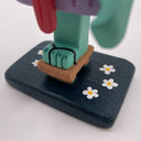 Close up of the base of a wooden sculpture, showing a teal colored foot wearing a wooden sandal on a black base with white daisies. 