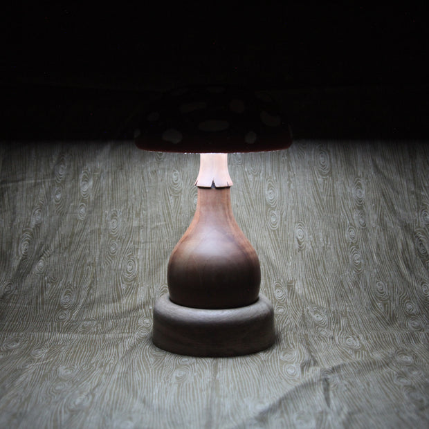 Carved wooden mushroom, with a thick and smooth wooden base. The top of the mushroom is red with while polka dots. Sculpture is also a lamp, emitting a dim glow.