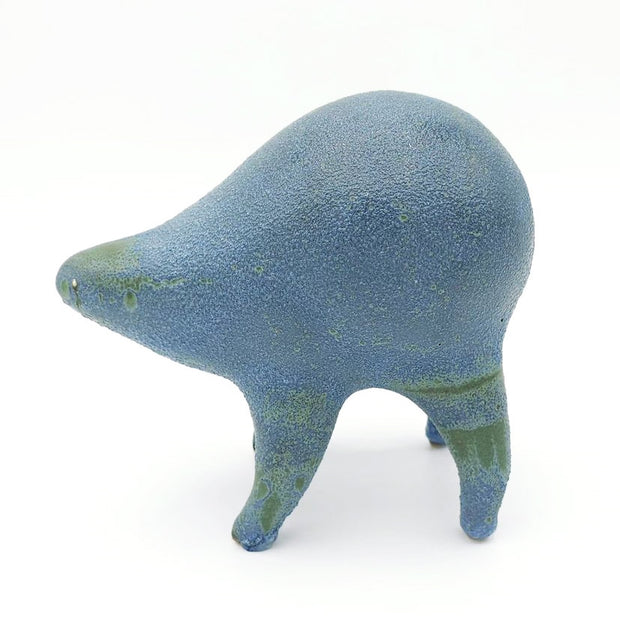 Greyish blue and green ceramic sculpture of a rounded body quadruped creature with a closed goofy smile. It has small golden eyes and a speckled texture on its body.