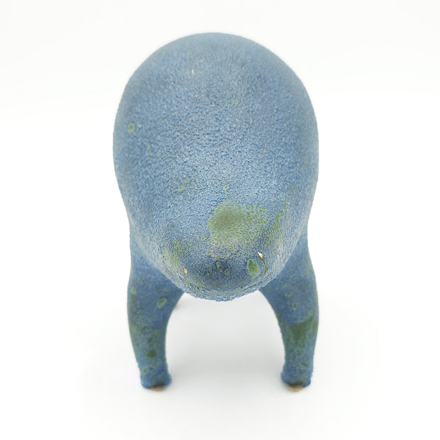 Greyish blue and green ceramic sculpture of a rounded body quadruped creature with a closed goofy smile. It has small golden eyes and a speckled texture on its body.