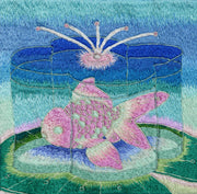 Embroidery of a pink and green goldfish inside of a flower shaped container of water. It sits atop a green lily pad with a blue gradient background.