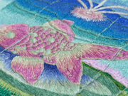 Embroidery of a pink and green goldfish inside of a flower shaped container of water. It sits atop a green lily pad with a blue gradient background.