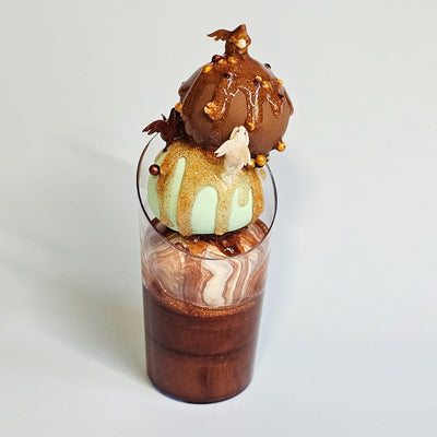 Sculpture of a triple scoop sundae in a cup of chocolate, mint and swirl. Sparkly sauce drips down it and nuts and small winged creatures adorn it. 