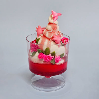 Sculpture of a ice cream sundae in a cup with strawberry syrup. It has small sculpted roses that decorate it and small pink winged creatures.