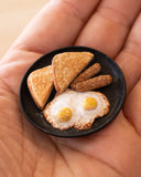 Small assembled clay sculpture of a breakfast on a plate of eggs, toast and 2 links of sausage. Held in the hand of the artist.